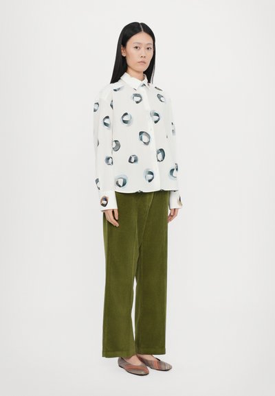 Camisa blanca con patrones circulares en azul y negro, combinada con pantalones de pana verde oliva. El modelo lleva zapatos planos a rayas multicolores.