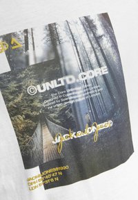 Hvid bomulds T-shirt med et grafisk print af en skov og en bro, med guld- og sort tekst inklusive brandnavn og koordinater.