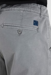 Pantalon en coton gris avec une poche arrière dotée d'une fermeture à bouton et d'une étiquette en tissu bleue portant l'inscription "DNM WRKRS". Texture lisse.