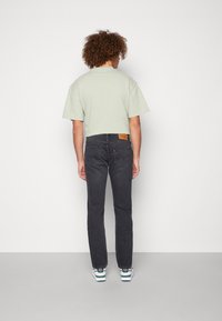 Persona con pelo rizado de espaldas, vistiendo una camiseta corta verde claro, vaqueros negros y zapatillas blancas sobre un fondo liso.