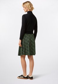 Schwarzer Rollkragenpullover, grüner Rock mit geometrischem Muster und schwarze Sneaker mit beigen Akzenten. Die Rückansicht hebt die Proportionen der Kleidung hervor.