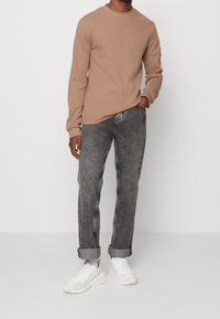 Beige Langarmhemd mit texturierter Oberfläche, kombiniert mit hellgrauen schmalen Jeans und weißen Sneakern mit Mesh-Details.