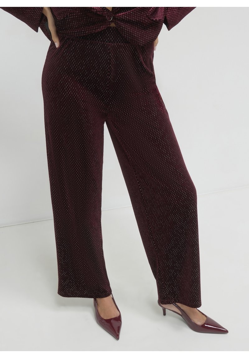 Femme portant un pantalon bordeaux à jambes larges texturé avec des talons slingback assortis à bout pointu sur un sol blanc uni.