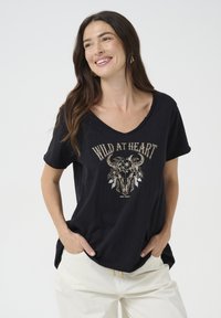 T-shirt noir à manches courtes avec un col en V, ornée d'un graphique central d'un crâne de taureau décoré de fleurs, et le texte "WILD AT HEART" au-dessus.