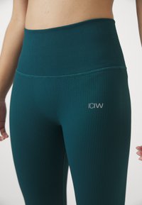 Cyan ribbellegging met een hoge tailleband en het logo "ICIW" in zilver. Het materiaal is rekbaar en zorgt voor een strakke pasvorm.