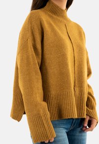 Pull en maille oversized jaune moutarde avec un col montant à côtes et des poignets larges à côtes, présentant un motif texturé subtil et un détail de couture centrale.