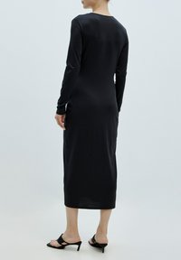 Schwarzes langärmliges Kleid aus einem glatten, dehnbaren Stoff, mit einer figurbetonten Silhouette und knielangem Saum. Kombiniert mit schwarzen Absatzsandalen.
