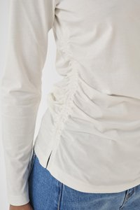 Off-white long-sleeve top with ruched detailing on the side worn with blue denim jeans, showing upper arm and waist area.