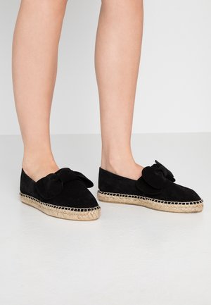 Jambes portant des espadrilles en suède noir avec des semelles en jute tissé et de grands nœuds décoratifs sur le dessus, posées sur un sol blanc lisse.