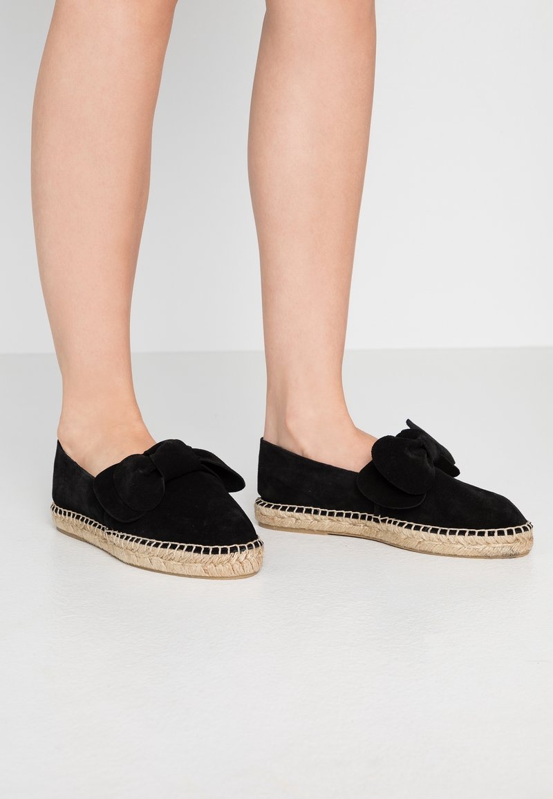 Jambes portant des espadrilles en suède noir avec des semelles en jute tissé et de grands nœuds décoratifs sur le dessus, posées sur un sol blanc lisse.