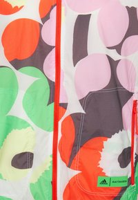 Tissu floral coloré avec de grandes formes de fleurs abstraites orange, vertes, roses et brunes, accompagné d'une fermeture éclair rouge, présentant les logos Adidas et Marimekko.