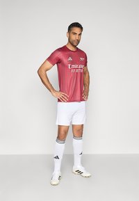 Rood adidas voetbalshirt met verticale strepen, witte korte broek en voetbalkousen met zwarte strepen, aangevuld met witte en zwarte voetbalschoenen.