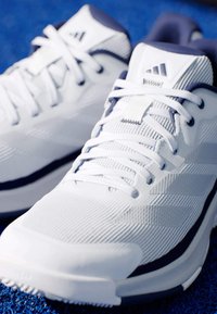 Bílé sportovní boty s texturovanou tkaninou, polstrovaným límcem a bílými šňůrkami. Mají námořnický akcent podél podrážky a pruhované designové prvky.