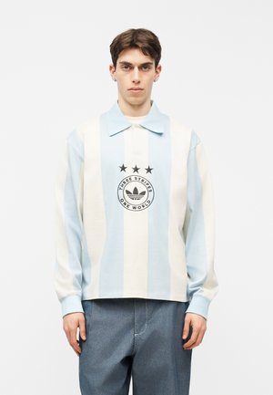 Bărbat tânăr purtând un tricou polo cu mânecă lungă, în dungi verticale albastre deschis și albe, cu un logo negru și textul „Three Stripes One World”.