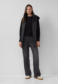 Zwarte puffer vest met rits, gedragen over een zwart geribbeld sweater, gecombineerd met donkergrijze wijde jeans en sneakers met beige accenten.