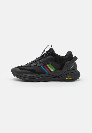 Czarny sneaker z wielokolorowymi paskami po boku oraz żółtym napisem "NEVER ASSUME" na podeszwie, blisko pięty.