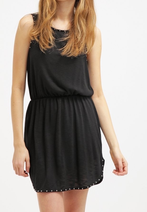 Femme portant une robe noire sans manches avec de petites clous argentés le long de l'encolure et de l'ourlet, debout devant un fond blanc uni.