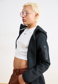 Un sweat à capuche noir zippé au design ajusté, avec une poche latérale zippée. Porté par-dessus un crop top blanc et un pantalon marron, avec des lunettes de soleil roses.