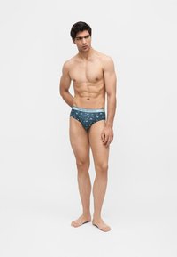 Άνδρας χωρίς πουκάμισο, όρθιος ξυπόλυτος, φορά μπλε εσώρουχα με σχέδια και ανοιχτό μπλε ζώνη, κοιτάζει στο πλάι σε απλό φόντο.