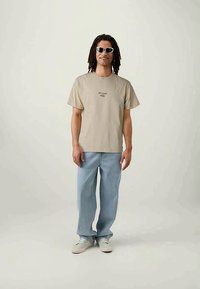 Beige Baumwoll-T-Shirt mit kleinem schwarzem Logo, kombiniert mit hellblauen Jeanshosen mit einem weißen Streifen und weißen Sneakern.