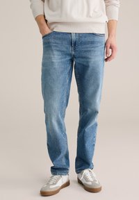 Šviesiai mėlynos spalvos džinsai tiesiu kirpimu, penkių kišenių dizainas ir subtilus išblukimas. Dėvimi su baltais sportbačiais su pilkomis detalėmis.