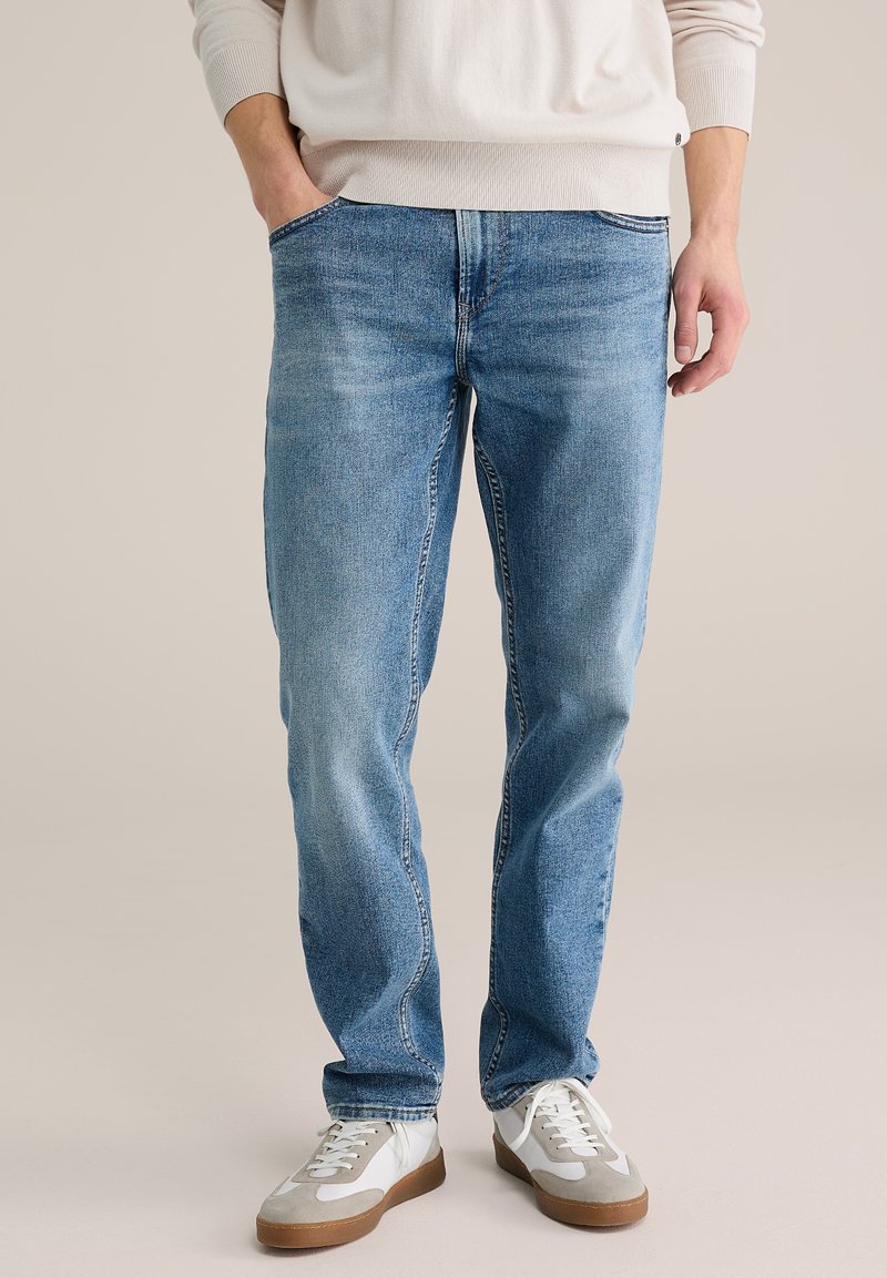 Šviesiai mėlynos spalvos džinsai tiesiu kirpimu, penkių kišenių dizainas ir subtilus išblukimas. Dėvimi su baltais sportbačiais su pilkomis detalėmis.