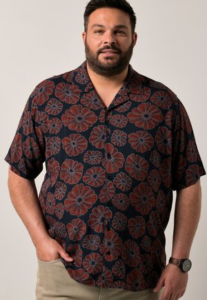 Mann trägt ein kurzärmeliges dunkles Hemd mit roten und blauen Blumenmustern, beige Hosen und eine Armbanduhr am linken Handgelenk.