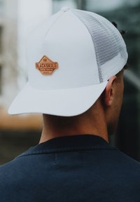 Hvid kasket med flad skygge, mesh-sider og et brunt læderpatch med teksten "BLACKSKIES PREMIUM SUPPLIES MMIX" på fronten.