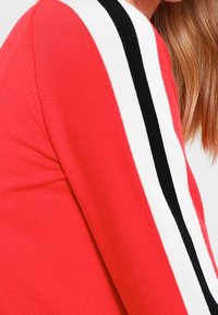 Pull rouge avec une texture côtelée. Présente des rayures verticales noires et blanches le long des manches. La vue rapprochée met en avant le matériau et le design.