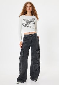 Valkoinen pitkähihaisuus crop-top, jossa on perhosgraafikka; yhdistetty mustiin cargo-housuihin, joissa on useita taskuja ja löysä istuvuus. Metallinväriset kengät.