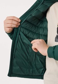 Main tenant une veste matelassée vert foncé ouverte, montrant une poche intérieure sur la doublure, sur un fond uni.