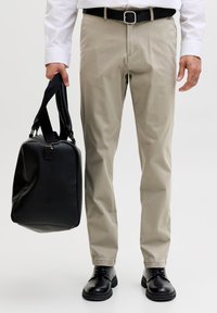 Mann iført beige bukser, svart belte, svarte sko, som holder en svart veske i håndtakene mot en ensfarget bakgrunn.