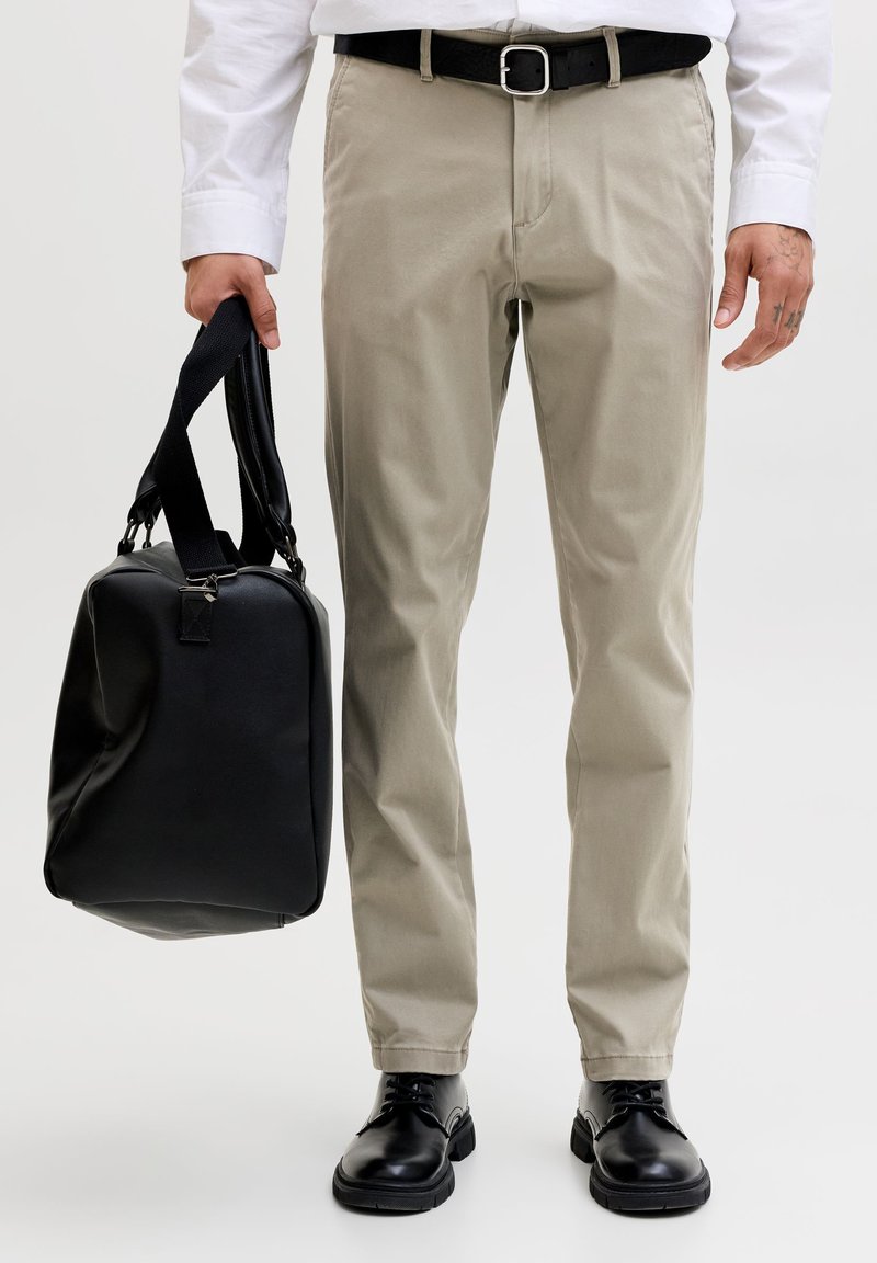 Mann iført beige bukser, svart belte, svarte sko, som holder en svart veske i håndtakene mot en ensfarget bakgrunn.