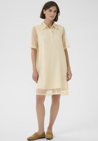 Helles gelbes Kleid mit Kragen und Knöpfen, Spitzenärmeln und einem Saum, der oberhalb des Knies endet. Weicher Stoff, lässiges Design.