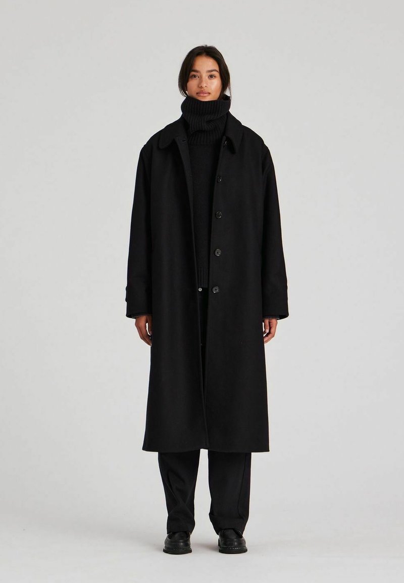 Sort lang frakke med bred krave, knaplukninger og en afslappet pasform. Båret over en matchende sort turtleneck og bukser i tekstureret stof.