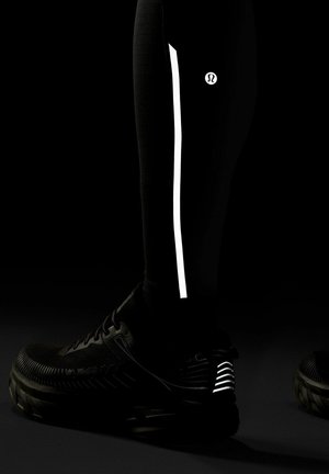 Alahasa ala betis yang mengenakan legging hitam dengan garis reflektif vertikal dan logo, dipadukan dengan sepatu lari hitam yang memiliki aksen reflektif di tumit.