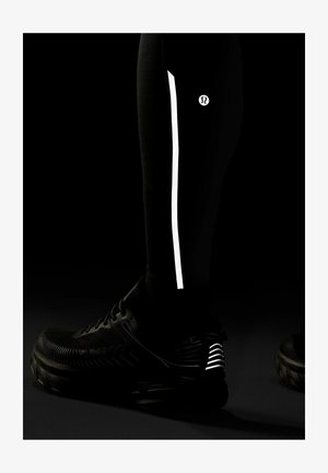 Alahasa ala betis yang mengenakan legging hitam dengan garis reflektif vertikal dan logo, dipadukan dengan sepatu lari hitam yang memiliki aksen reflektif di tumit.