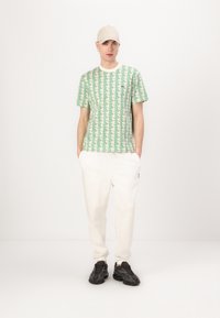 Tricou scurt cu mânecă, modelat verde, pantaloni jogger albi și adidași negri. Tricoul are un imprimeu geometric pe un fundal deschis.