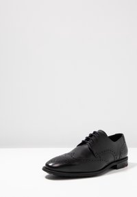 Chaussure habillée en cuir noir avec un design en pointe d'aile, des détails perforés, un bout rond, et une fermeture à lacets à cinq œillets. Semelle plate avec des accents subtils.