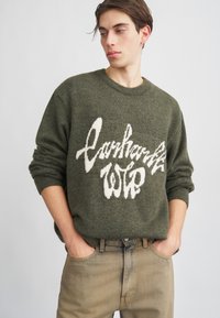 Pulover tricotat de culoare verde măsliniu, cu un design alb al logo-ului pe față, guler rotund și manșete cu model, purtat cu blugi din denim bej deschis.