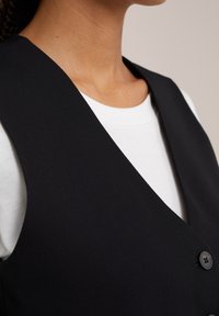 Czarna kamizelka z dekoltem w serek, wykonana z gładkiego materiału. Posiada jeden czarny guzik i jest noszona na białej koszulce.