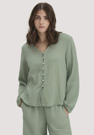 Frau mit schulterlangem braunem Haar, trägt ein hellgrünes, strukturiertes Button-up-Oberteil und passende Hose, Hände in den Taschen, neutrale Hintergrund.