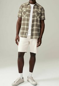 Muž v béžové kostkované krátké košili přes bílé tričko, krémové kraťasy, bílé ponožky a béžové tenisky stojí před jednobarevným pozadím.