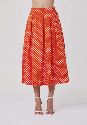 Femme portant une jupe midi orange taille haute à pois blancs et des talons blancs à bride cheville, debout devant un fond uni.