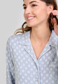 Mujer sonriente ajustando un pequeño pendiente de botón, vistiendo pijama azul claro con un patrón de hojas blancas y cuello con ribete.