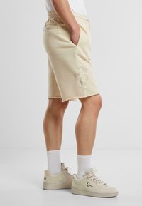 Beige kokvilnas šorti ar relaksētu piegriezumu, ar dekoratīvām izšuvuma detaļām un saplēstu apakšmalu, kopā ar beige čībām un baltiem zeķēm.