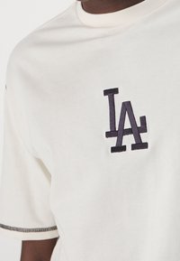 Krémové bavlněné tričko s výšivkou navy "LA" na levé straně hrudi. Krátké rukávy a kontrastně šité lemování.