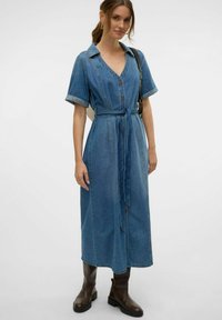 Denim haljina u svijetloplavoj boji, s V-izrezom, kratkim zavrnutim rukavima, dugmadi sprijeda, remenom u struku i bočnim džepovima. Uparena s smeškim čizmama.