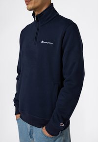 Uomo che indossa un felpa blu scuro Champion con zip a quarti e logo sul petto e sulla manica, con le mani nelle tasche, abbinata a jeans azzurri.