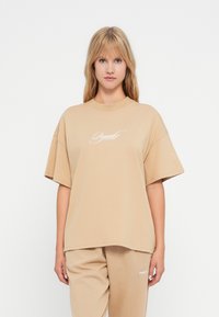 Žena oblečená v béžovém oversized tričku a odpovídajících kalhotách s bílým nápisem na tričku a kalhotách, stojící před bílým pozadím.